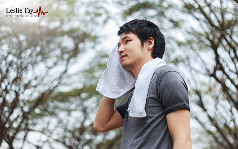 A man in a grey athletic shirt wipes sweat from his face with a white towel while standing outdoors among trees.
